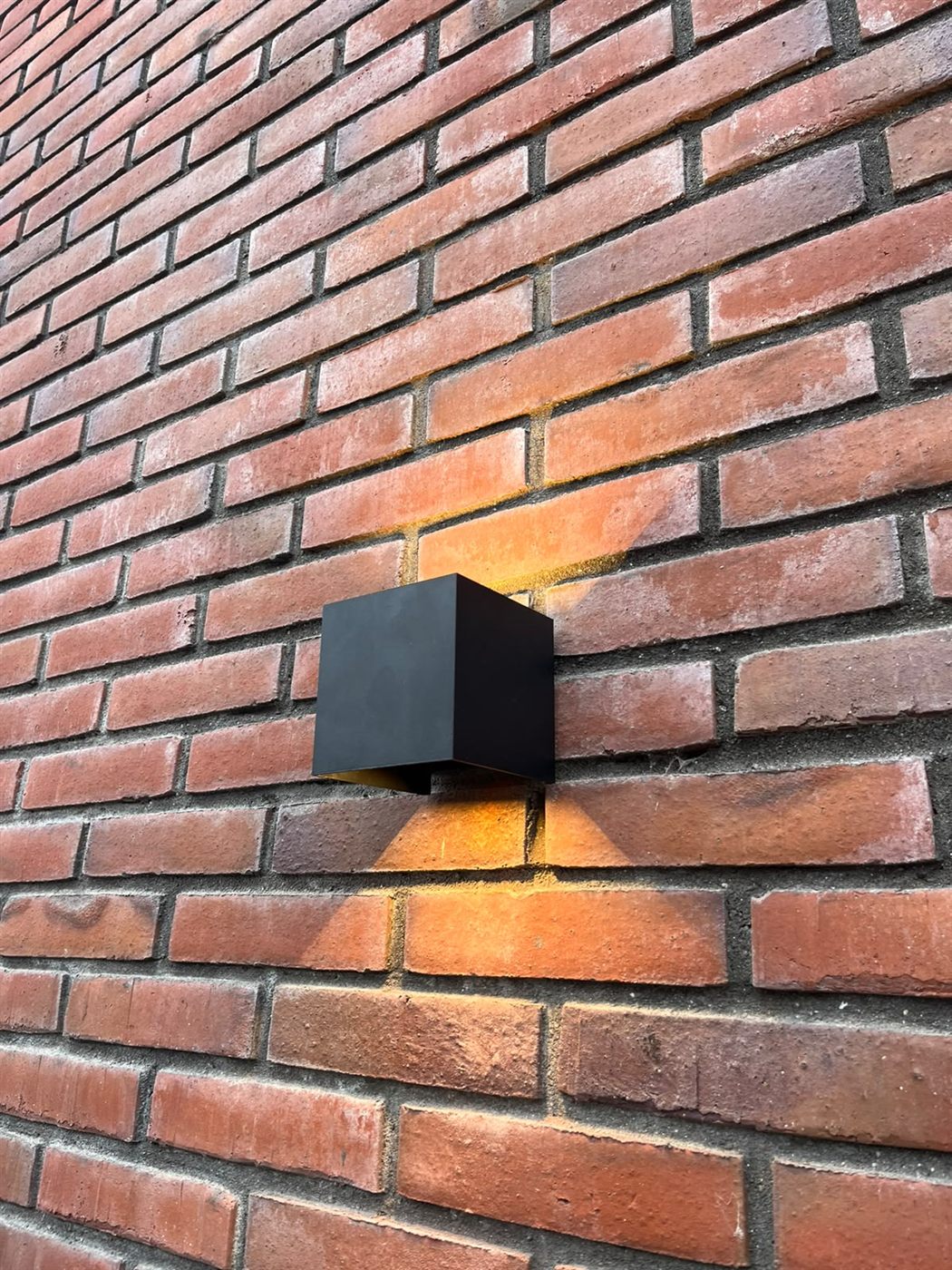 Zwarte kubus-wandlamp met warme up- en downlight op een rode bakstenen muur